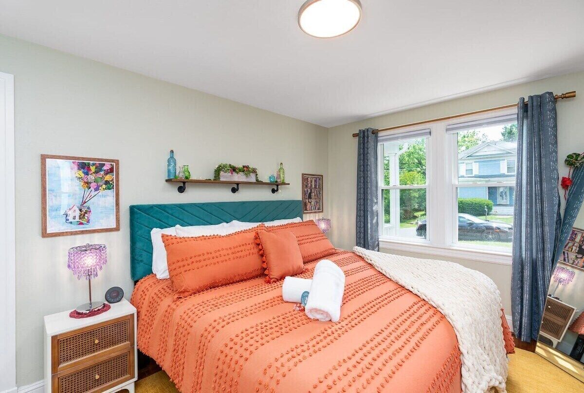 Un dormitorio con cabecera color esmeralda, cama con ropa de cama naranja, mesita de noche con lámpara y ventana con cortinas.