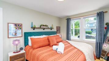 Un dormitorio con cabecera color esmeralda, cama con ropa de cama naranja, mesita de noche con lámpara y ventana con cortinas.