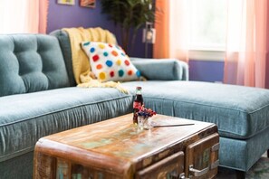Una sala de estar con un sofá azul, un cojín de lunares coloridos y una maleta antigua sobre una mesa de centro de madera.