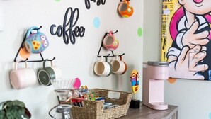 A wall with mugs hanging on a rack, a basket with snacks, and a coffee machine on a wooden table.
