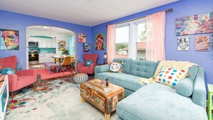 A vibrant living room with a blue sofa, a red armchair, and a coffee table. There is a kitchen visible through an archway.