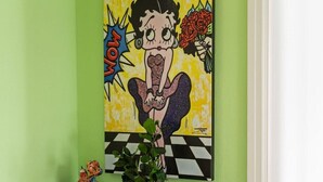 Ein Zimmer mit grüner Wand, einem Bild einer Frau, einem kleinen Tisch mit Büchern und einer Pflanze sowie einem Regal mit Handtüchern.