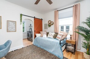 Ein Schlafzimmer mit einem Bett, einem Sessel, einem Teppich, einer Pflanze und einem Fenster mit Vorhängen.