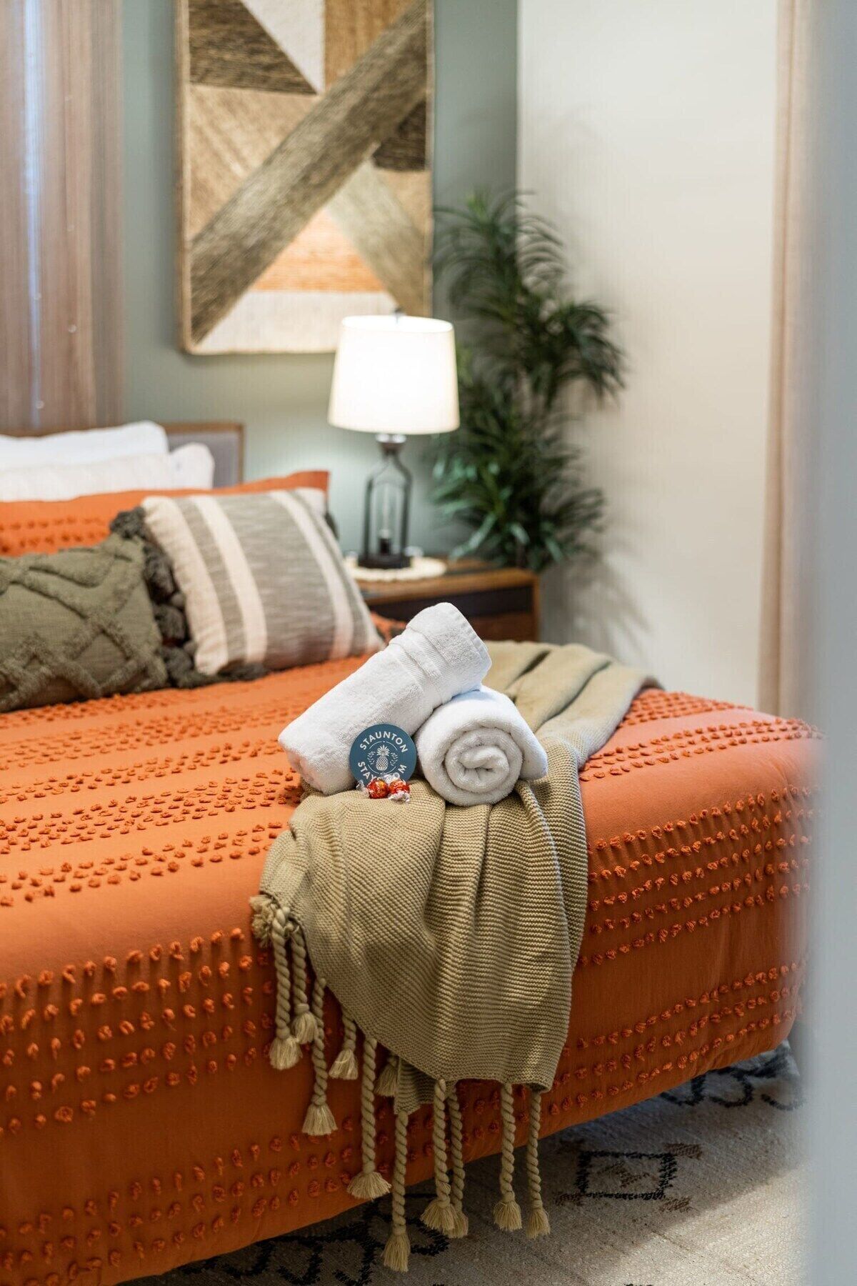 A neatly made bed with a rolled white towel and a blanket, a lamp, and a plant on a nightstand.