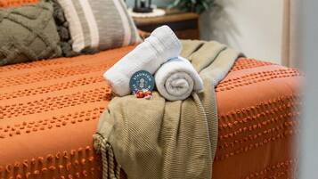A neatly made bed with a rolled white towel and a blanket, a lamp, and a plant on a nightstand.