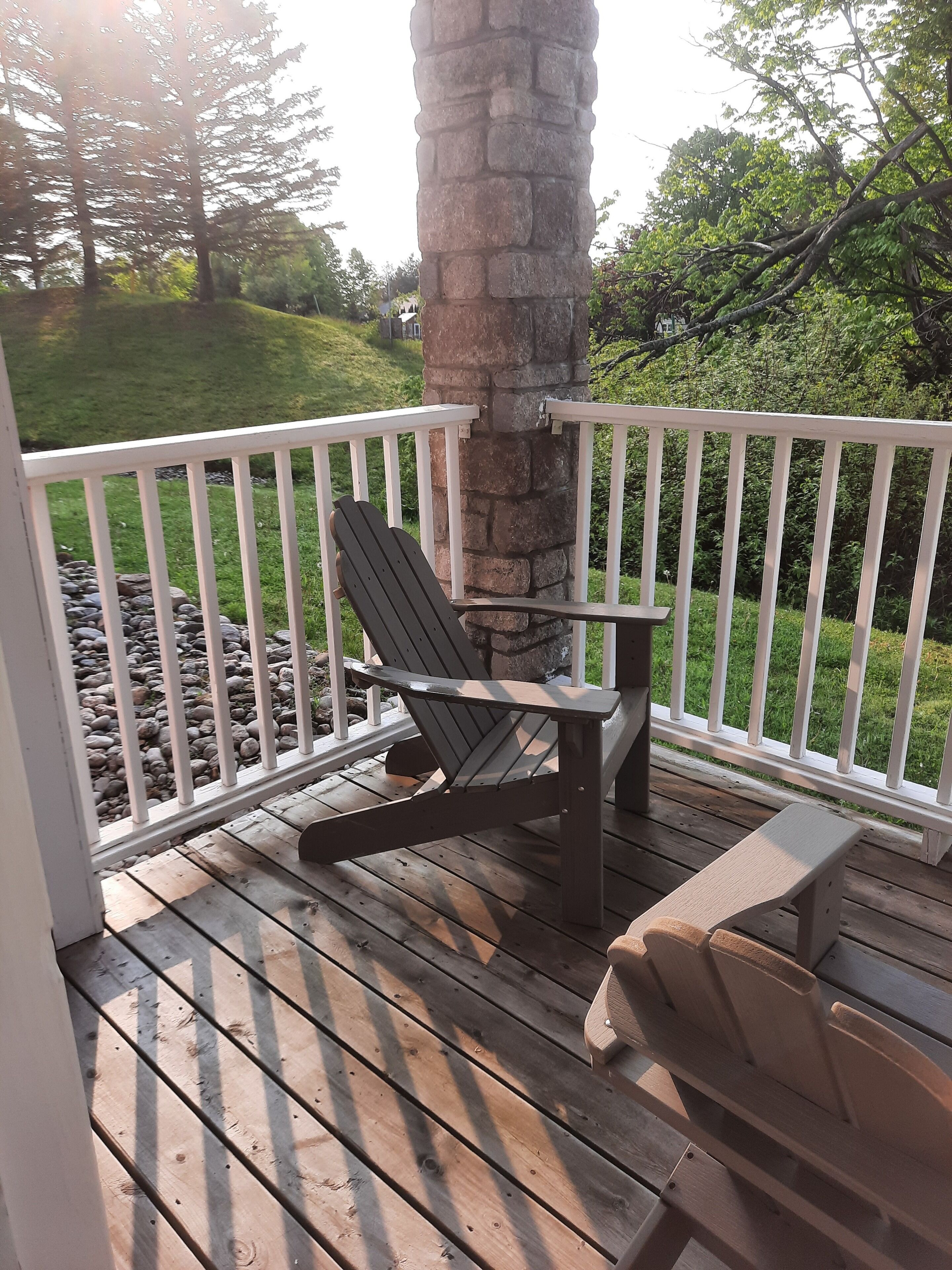 A wooden deck with a stone pillar, a white railing, and a single Adirondack chair.