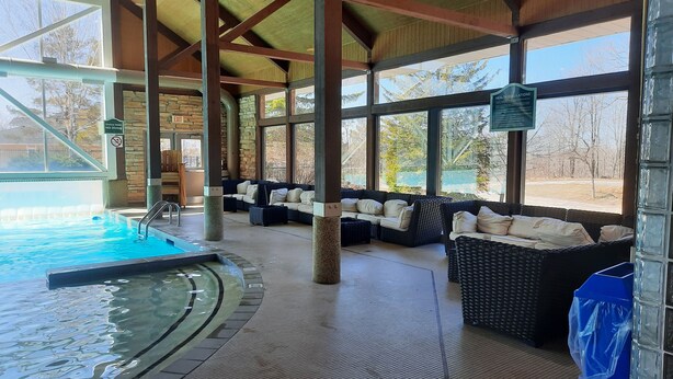 A covered indoor swimming pool area with a stone wall, a lounge with wicker furniture, and a view of trees outside.