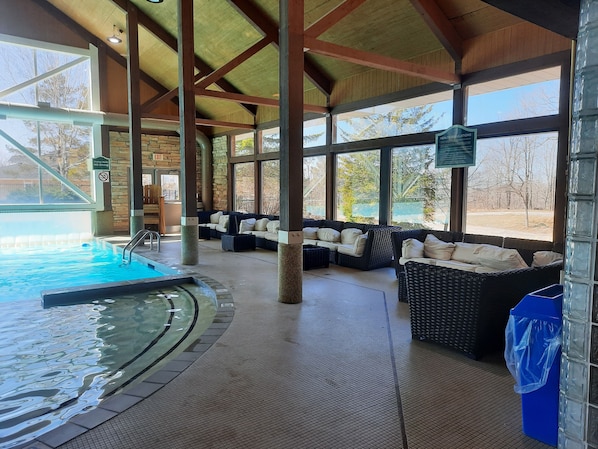 A covered indoor swimming pool area with a stone wall, a lounge with wicker furniture, and a view of trees outside. - Margarita Vacation Getaway (Oro-Medonte)