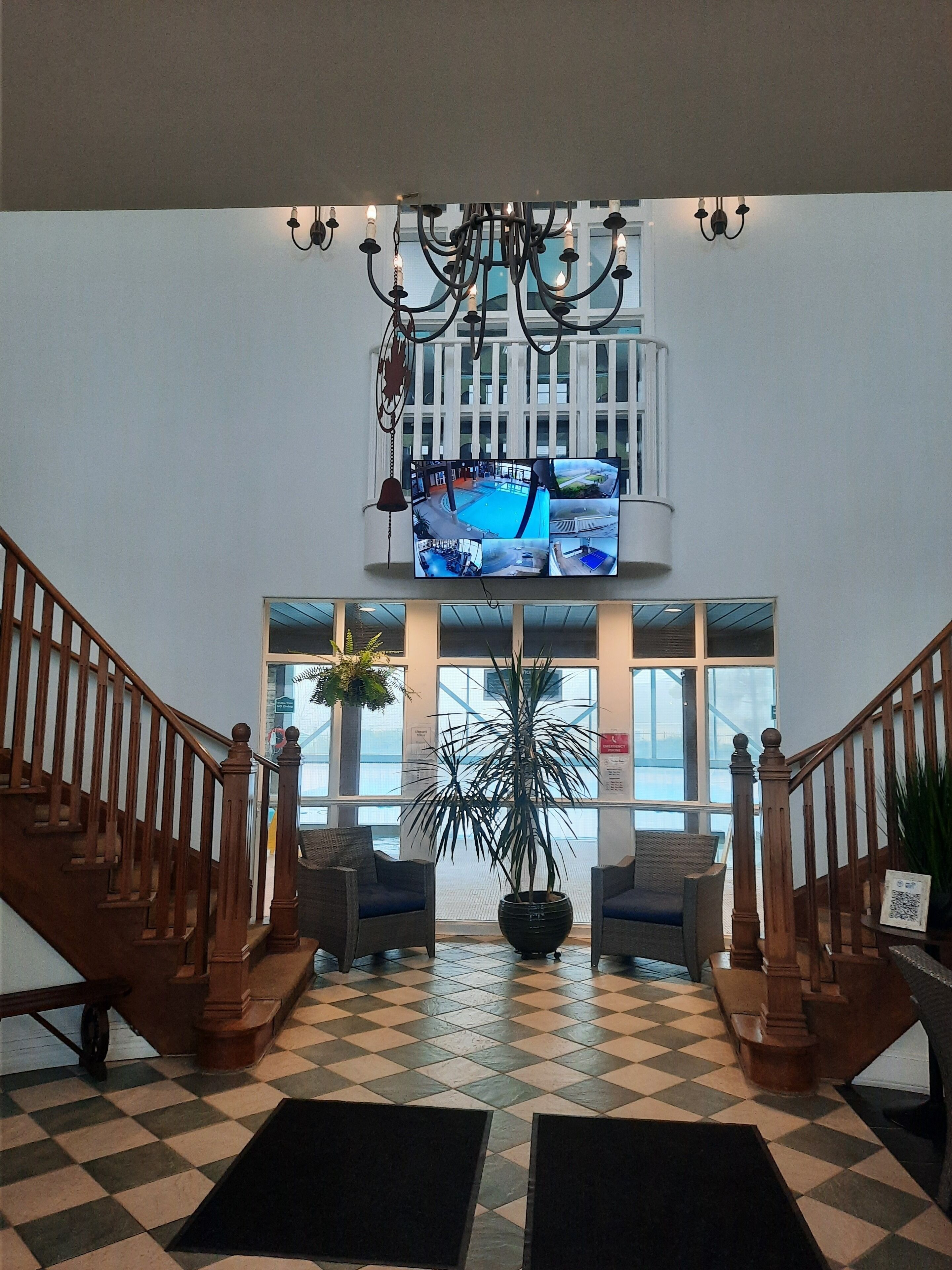Eine Lobby mit Schachbrettmuster-Boden, einer Holztreppe, einer großen Küstertopf-Pflanze und einem an der Wand befestigten Fernseher, der mehrere Ansichten zeigt.