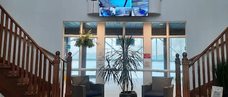 Een hal met een geruit vloerkleed, een houten trap, een grote potplant en een wandtelevisie die verschillende beelden toont.