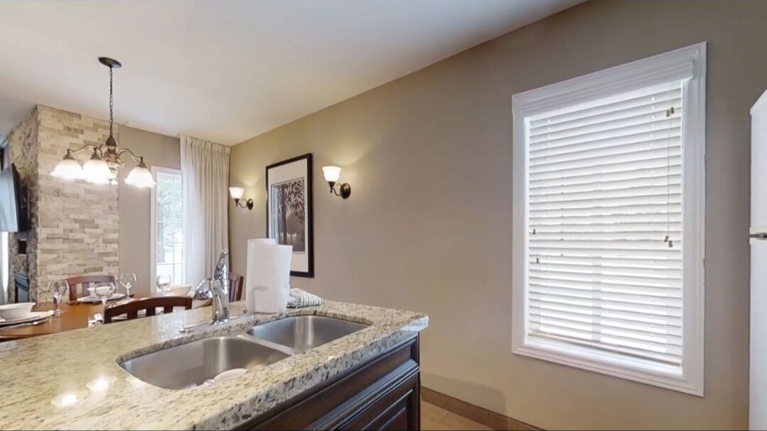 A kitchen with granite countertops, a double sink, a stone backsplash, a hanging light fixture, and a window with blinds.