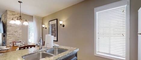 A kitchen with granite countertops, a double sink, a stone backsplash, a hanging light fixture, and a window with blinds.