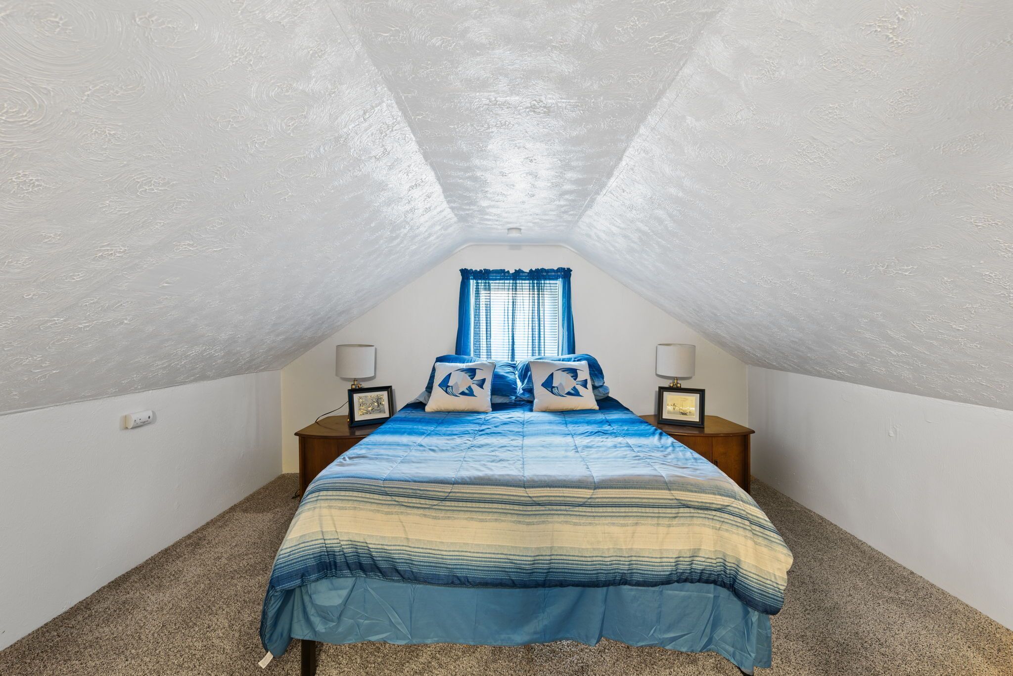 Une chambre avec un plafond en pente, un lit recouvert d’une couvre-lit à rayures bleues et blanches, deux oreillers, une table de chevet avec une lampe et une fenêtre avec des rideaux bleus.