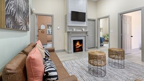A living room with a fireplace, a sofa, two wicker stools, and a flat-screen TV mounted above the fireplace. - Welcome to Casa Azul 1932 Eclectic Boho Sanctuary  in Downtown Riverside! (Riverside)