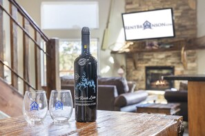 Una botella de vino y dos copas sobre una mesa de madera en una habitación con chimenea de piedra y un televisor al fondo.