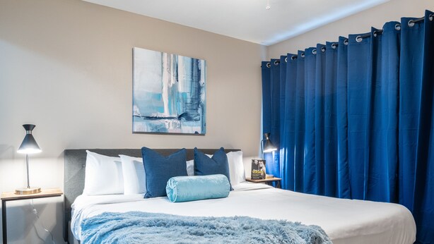 Un dormitorio con una cama grande, una mesita de noche con lámpara, un cuadro en la pared y cortinas azules.