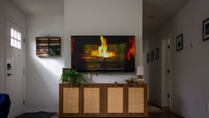A living room with a large flat-screen TV mounted on the wall, a wooden console table with a plant, and a framed welcome sign. - The Leland Blue on M-22 (Suttons Bay)