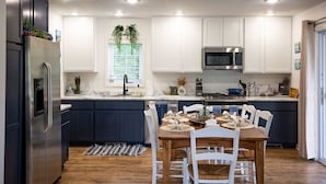 A modern kitchen with white cabinets, a stainless steel refrigerator, and a dining area with a wooden table and white chairs. - The Leland Blue on M-22 (Suttons Bay)