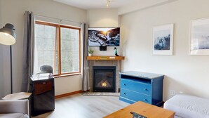 A modern living room with a fireplace, a television mounted above it, and a blue chest of drawers. - Hearthstone Lodge #417 (Sun Peaks)