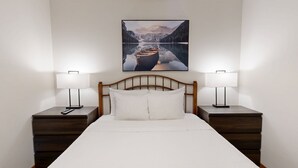 A neatly made bed with a wooden headboard, two bedside tables with lamps, and a framed picture of a boat on a lake above the bed. - Hearthstone Lodge #417 (Sun Peaks)