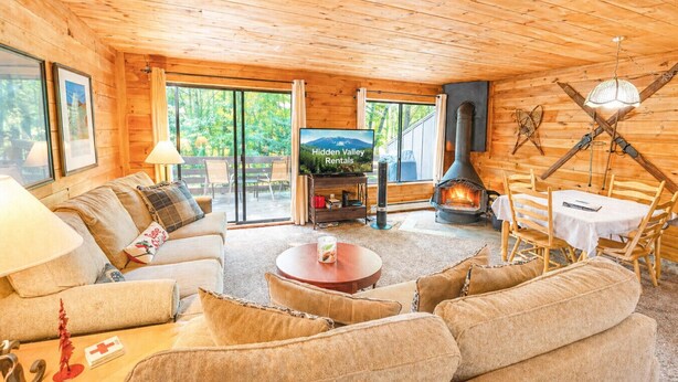 A cozy log cabin interior with a sofa, a television, a fireplace, and a dining area.