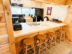 A wooden cabin interior with a kitchen, bar stools, and a dining table.