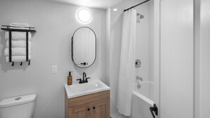 A bathroom with a toilet, sink, mirror, and shower. - Riverside Suite | Kitchenette, Firepit & BBQ (Lake Cowichan)