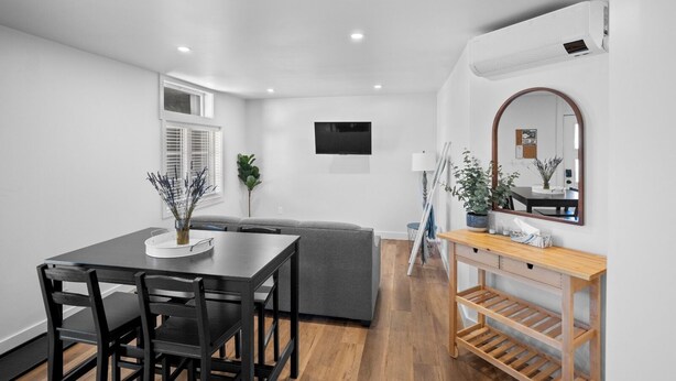 A modern living room with a dining table, grey sofa, wooden floor, and a TV mounted on the wall. - Riverside Suite | Kitchenette, Firepit & BBQ (Lake Cowichan)