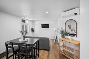 A modern living room with a dining table, grey sofa, wooden floor, and a TV mounted on the wall. - Riverside Suite | Kitchenette, Firepit & BBQ (Lake Cowichan)