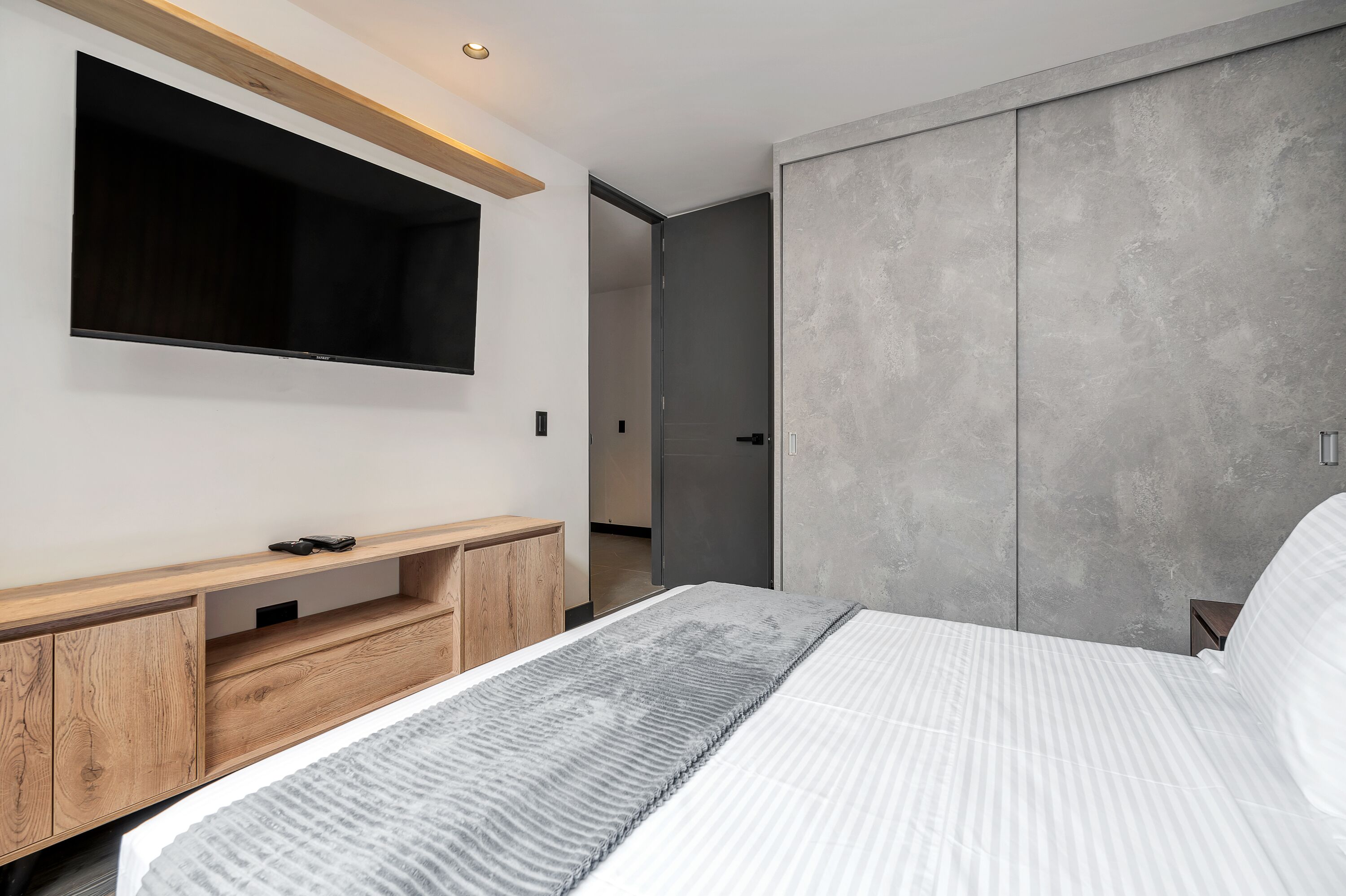 A modern bedroom with a bed, built-in TV, wooden storage unit, and a grey wardrobe.