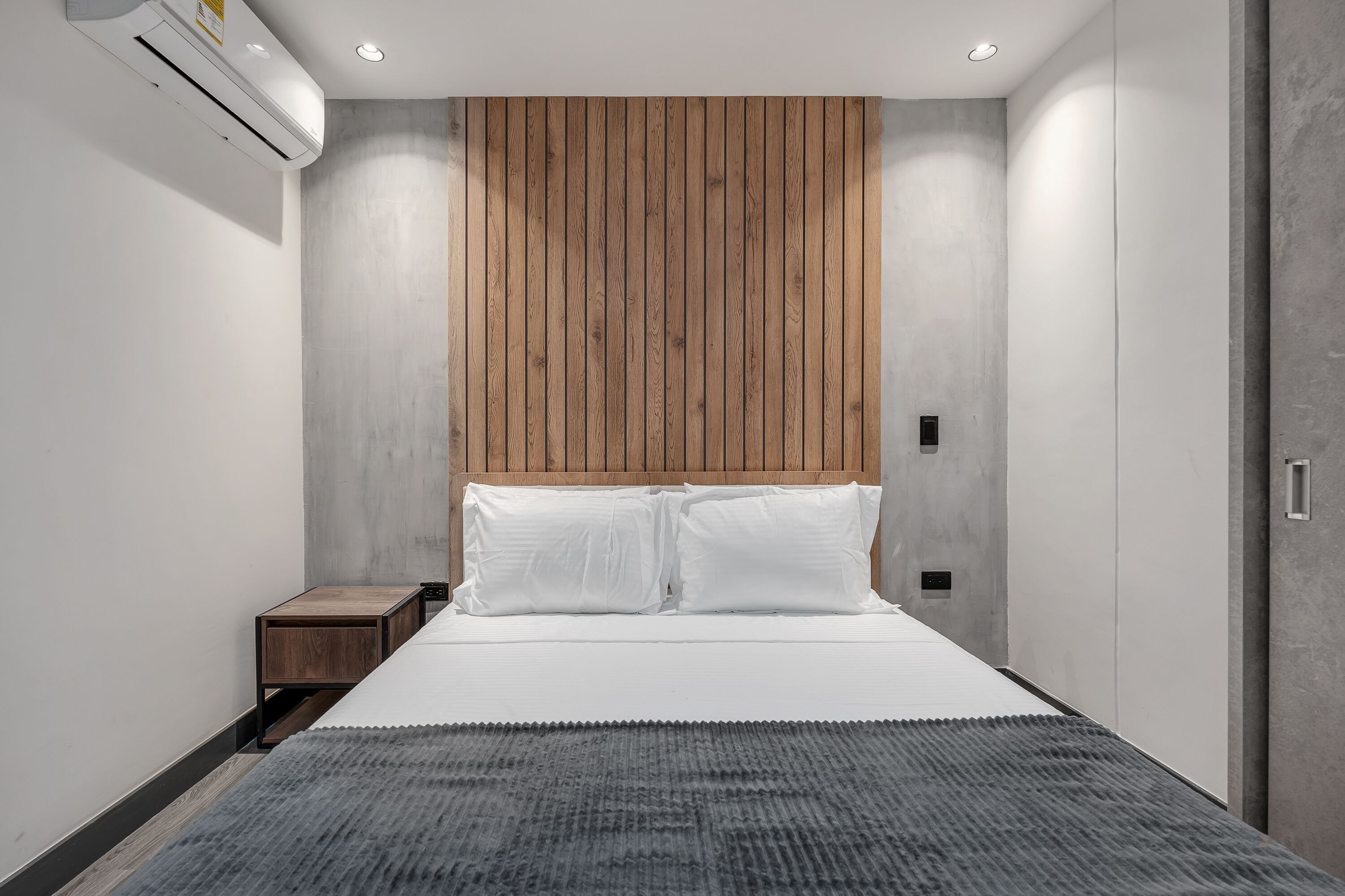 A modern bedroom with a wooden headboard, a bed with white linens, a small wooden nightstand, and a grey rug.