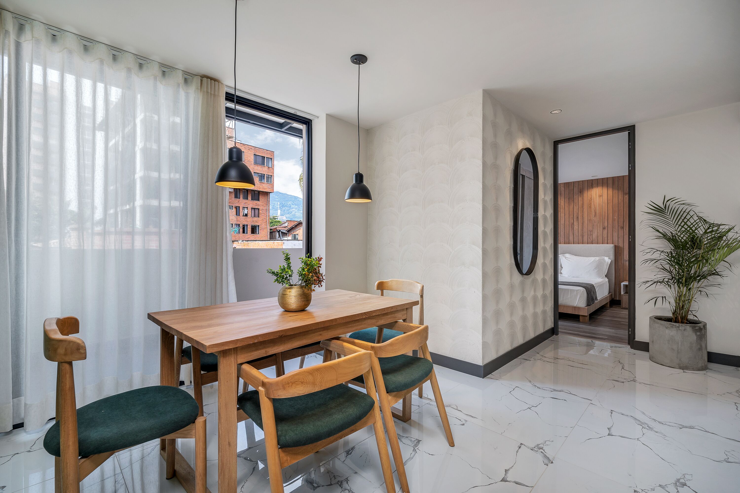 Habitación de hotel moderna con una mesa de comedor de madera, sillas con acolchado verde, una planta en maceta y vistas de un paisaje urbano a través de la ventana.