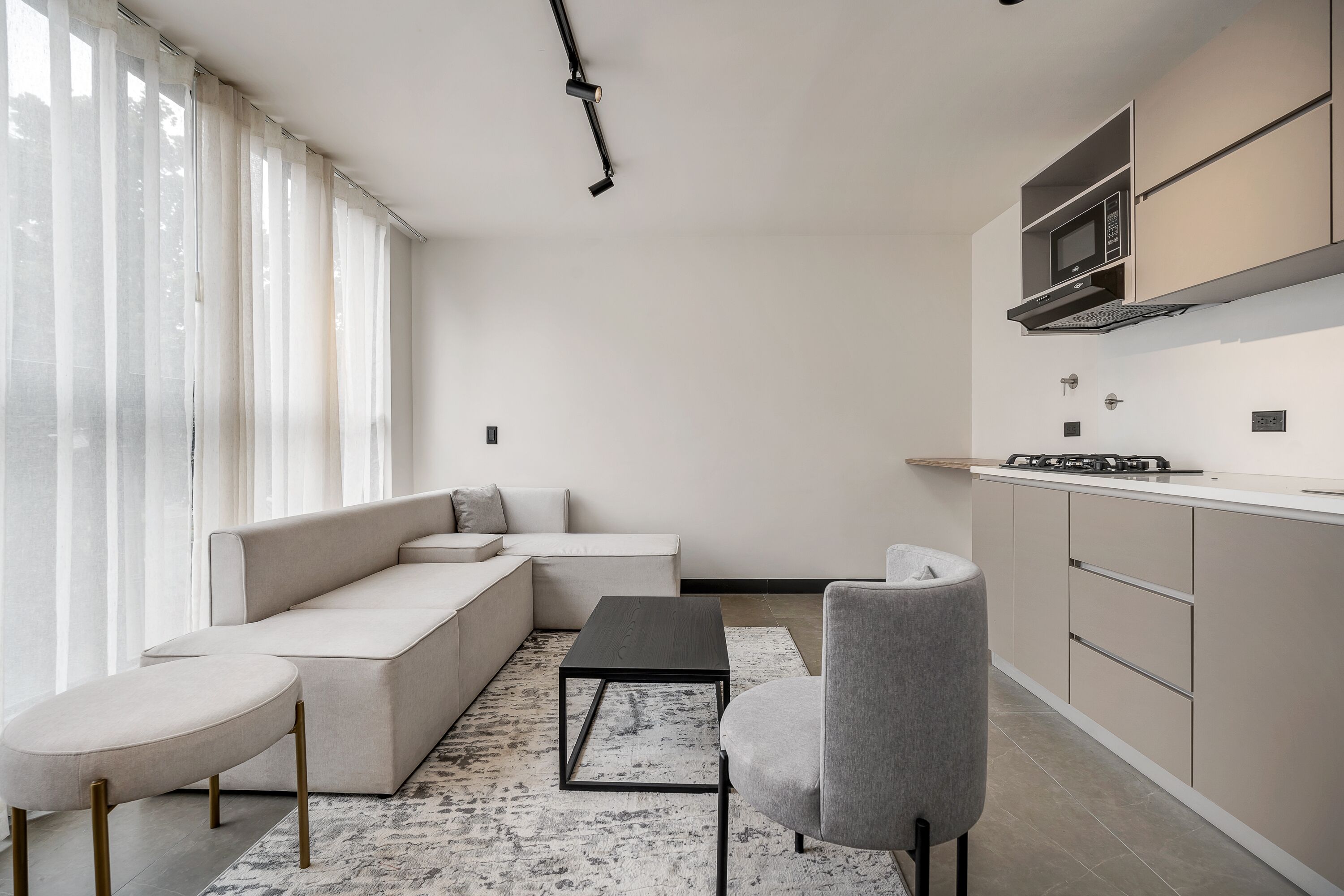 Ein modernes Wohnzimmer mit einer Couch, einem Couchtisch und einem Sessel. Es gibt eine Küchenzeile mit Schränken und einer Mikrowelle.
