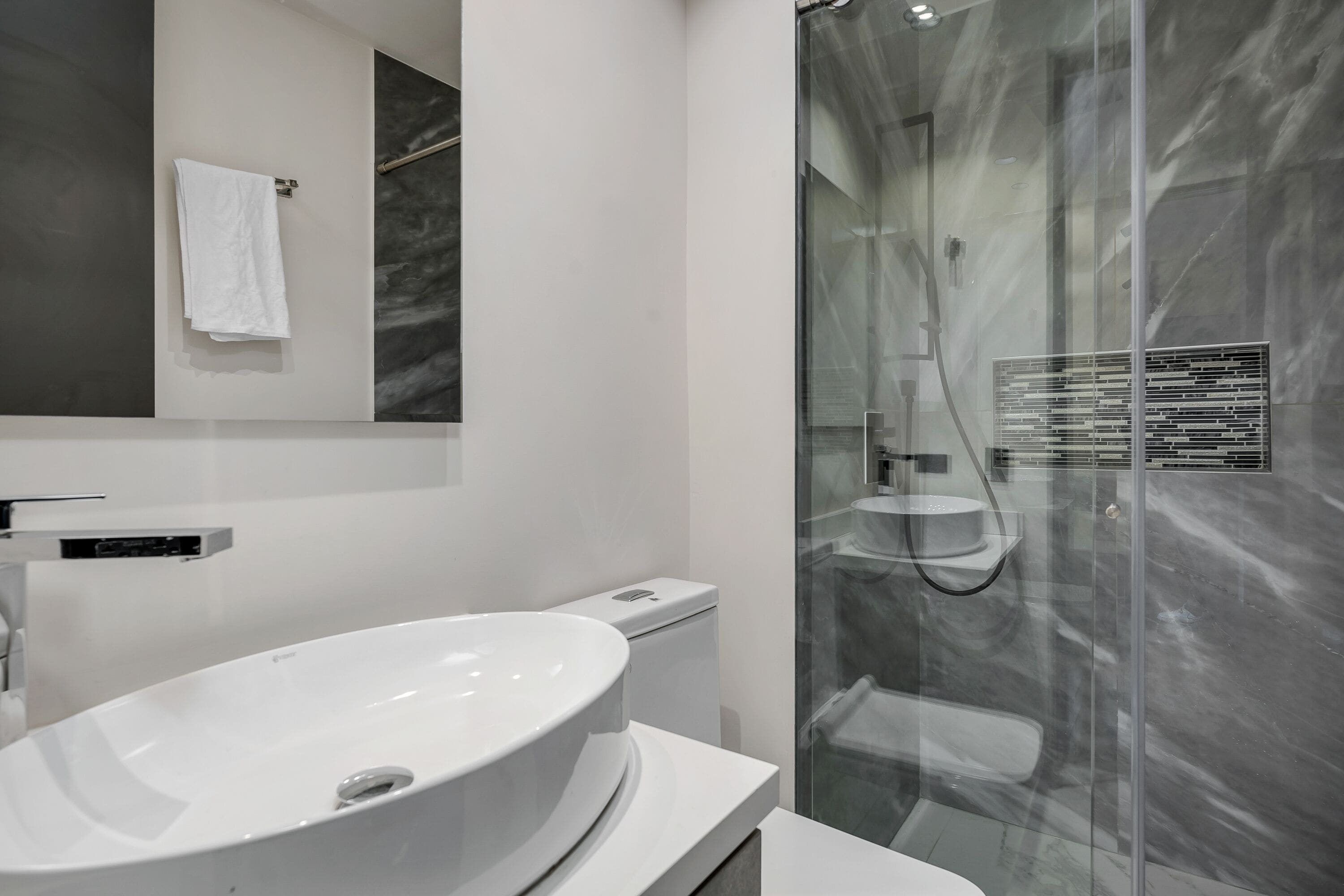 A modern bathroom with a large white sink, a glass-enclosed shower, and a marble wall.
