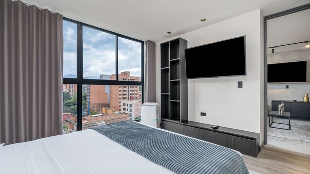 Une chambre d’hôtel moderne dotée d’un grand lit, d’un téléviseur fixé au mur et offrant une vue sur le paysage urbain grâce à de grandes fenêtres.