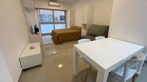 Una habitación pequeña y moderna con una cama, un escritorio y una silla. Se ve una ventana y unidades de aire acondicionado en el exterior.