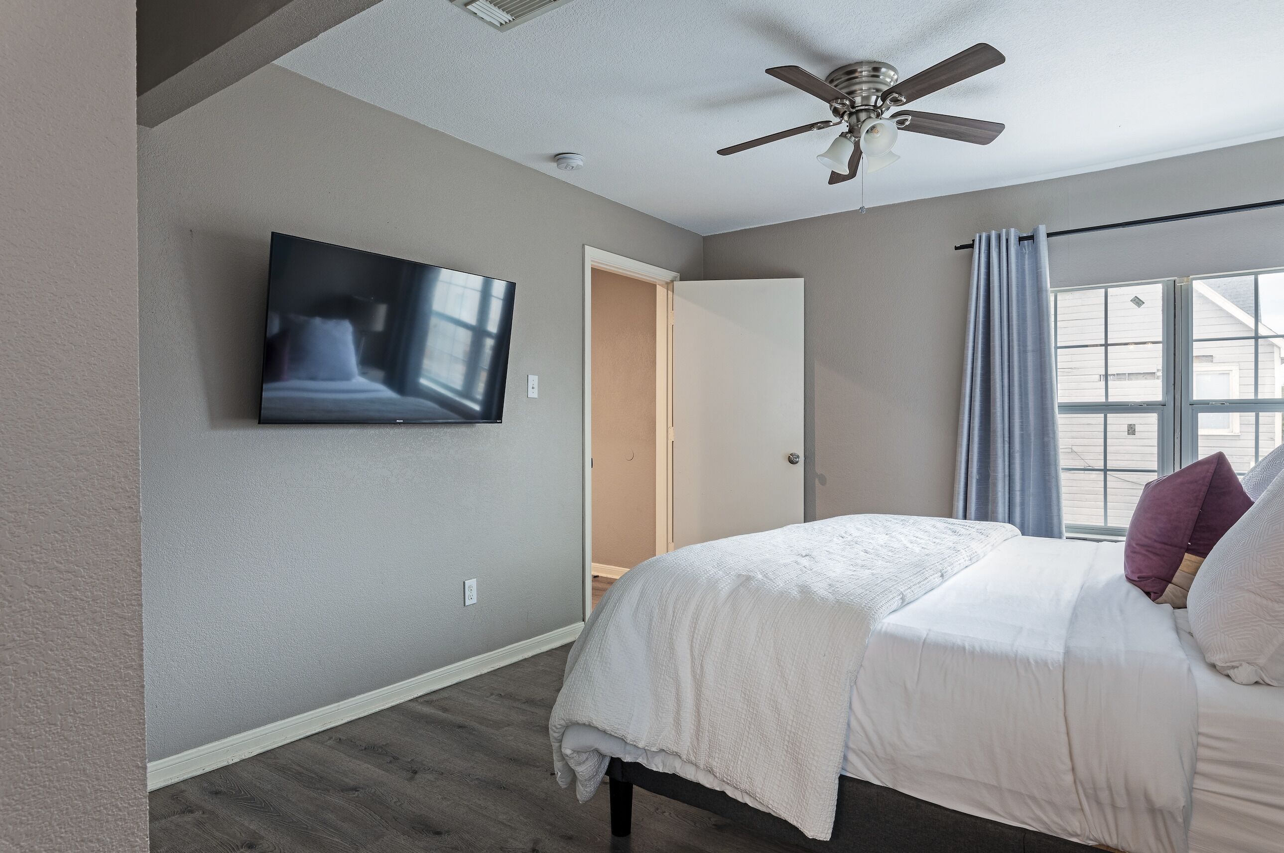 Une chambre à coucher avec un lit, un ventilateur de plafond, un téléviseur fixé au mur et une fenêtre avec des rideaux.