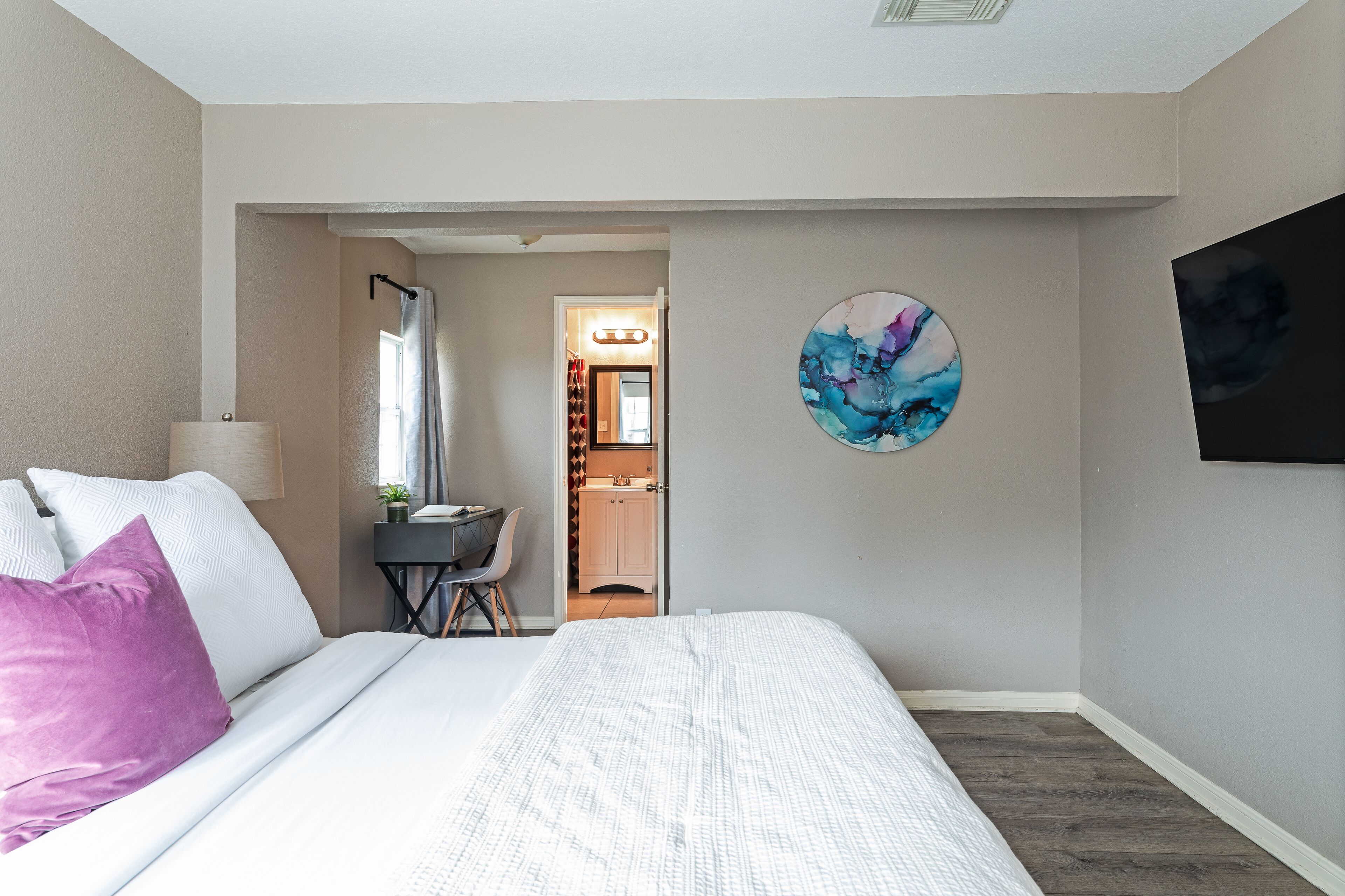 A bedroom with a bed, a desk, a chair, a TV, and a bathroom visible through an open door.
