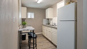 Una cocina compacta con encimera de mármol, armarios blancos, un refrigerador y una ventana con persianas.