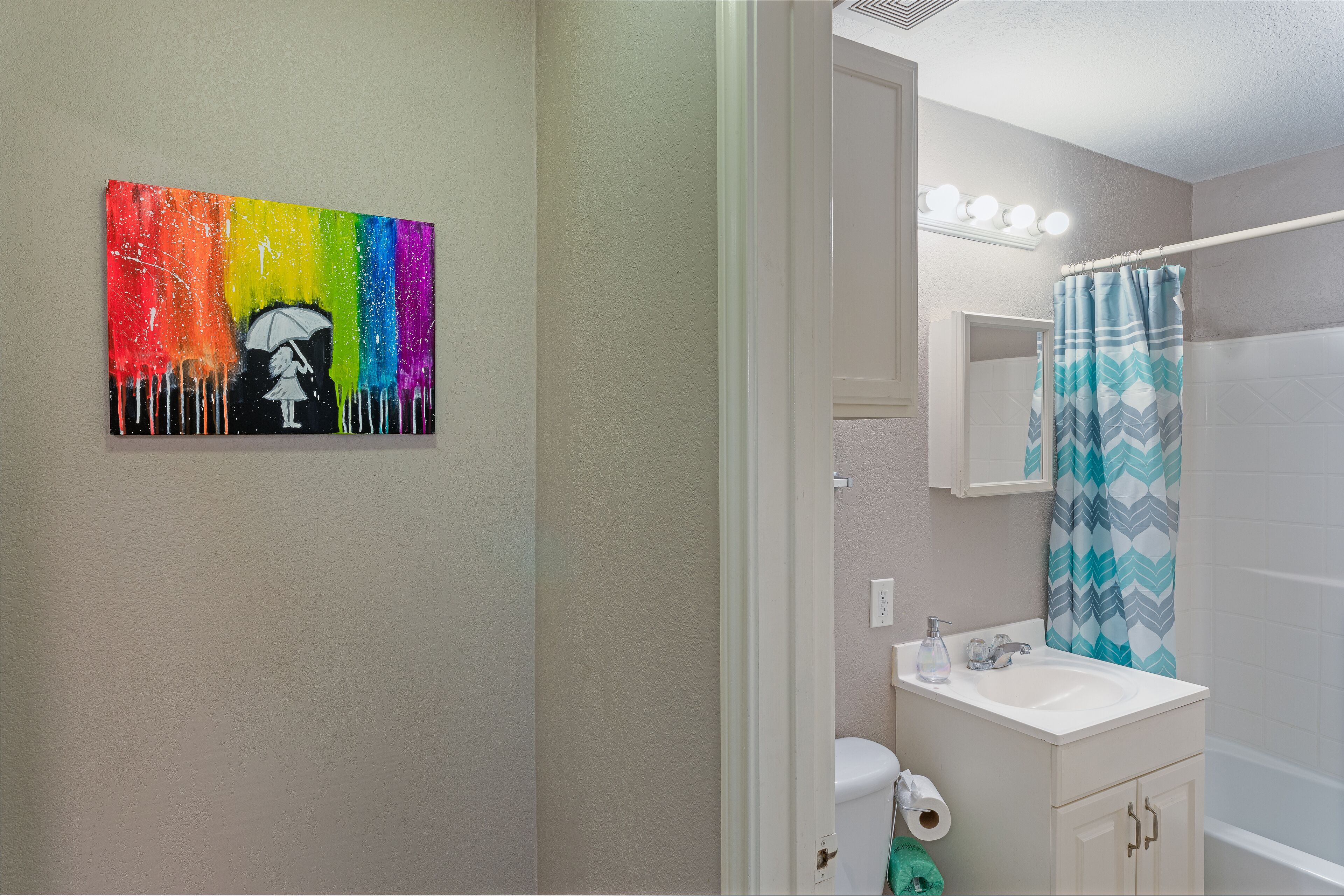 A bathroom with a white sink, a mirror, a shower curtain, and a painting on the wall.