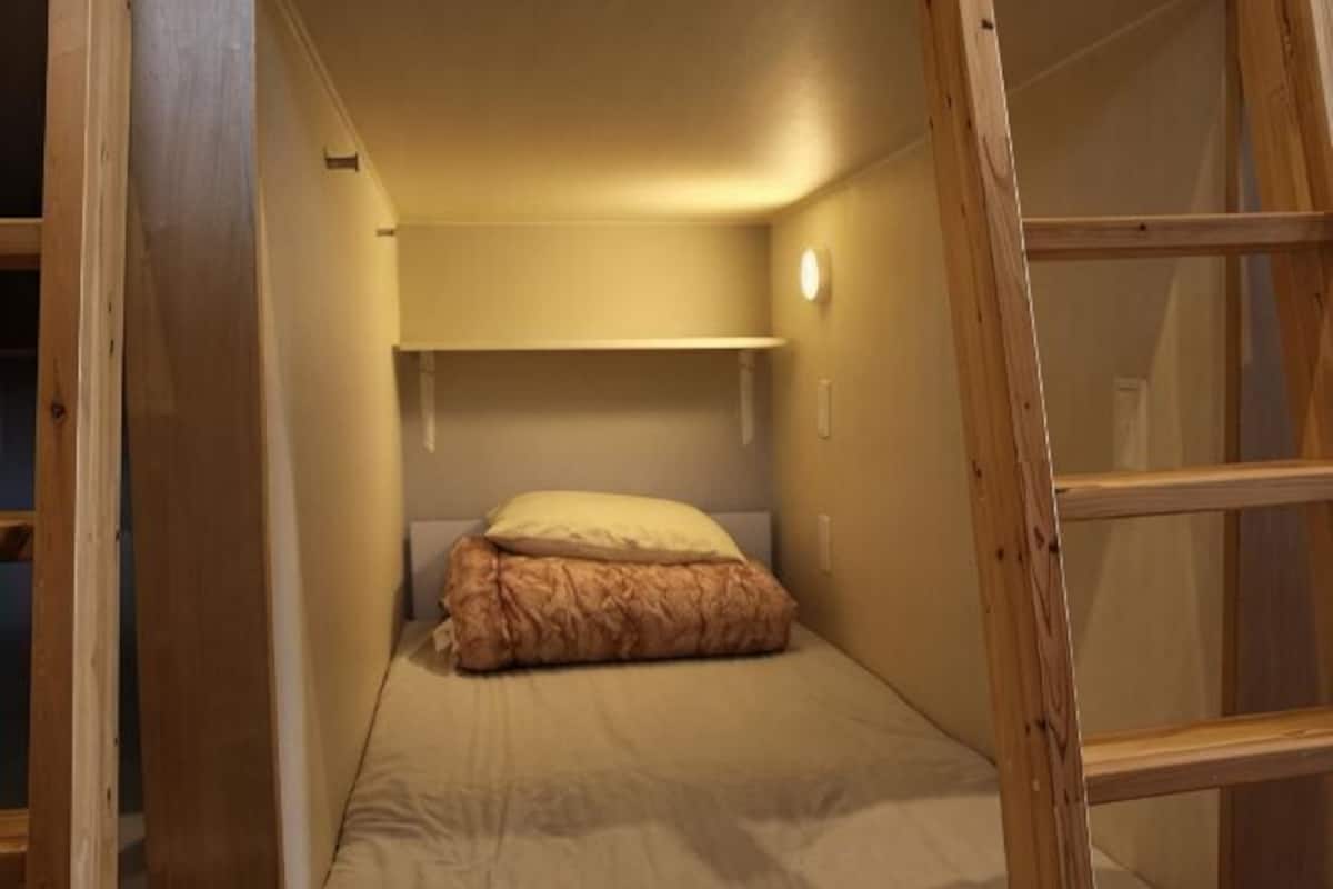 A narrow room with a single bed, a wooden ladder, and a shelf above the bed.