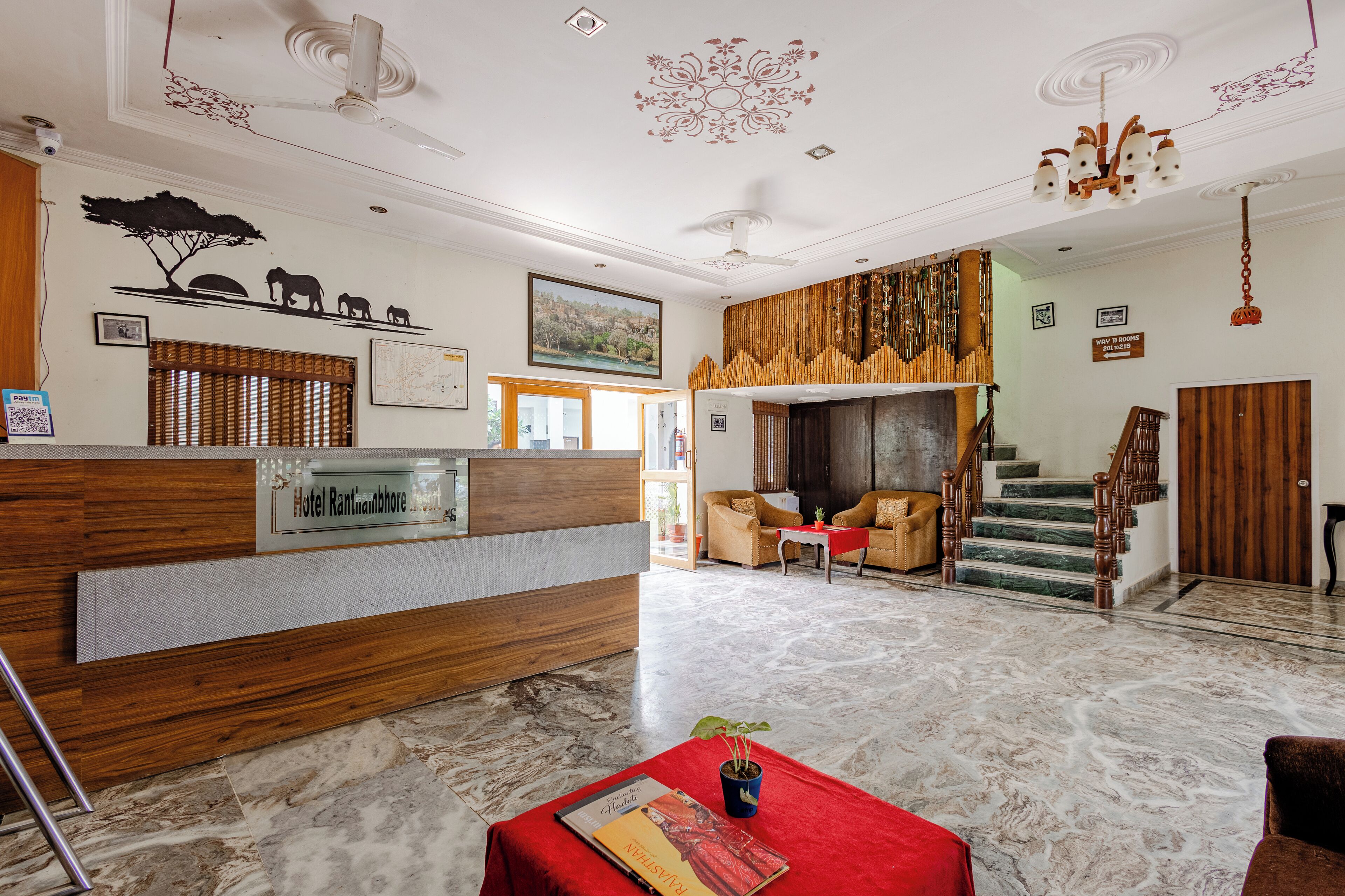 Vestíbulo de hotel con mostrador de recepción, área de estar, escalera y elementos decorativos como cuadros en la pared y un candelabro.
