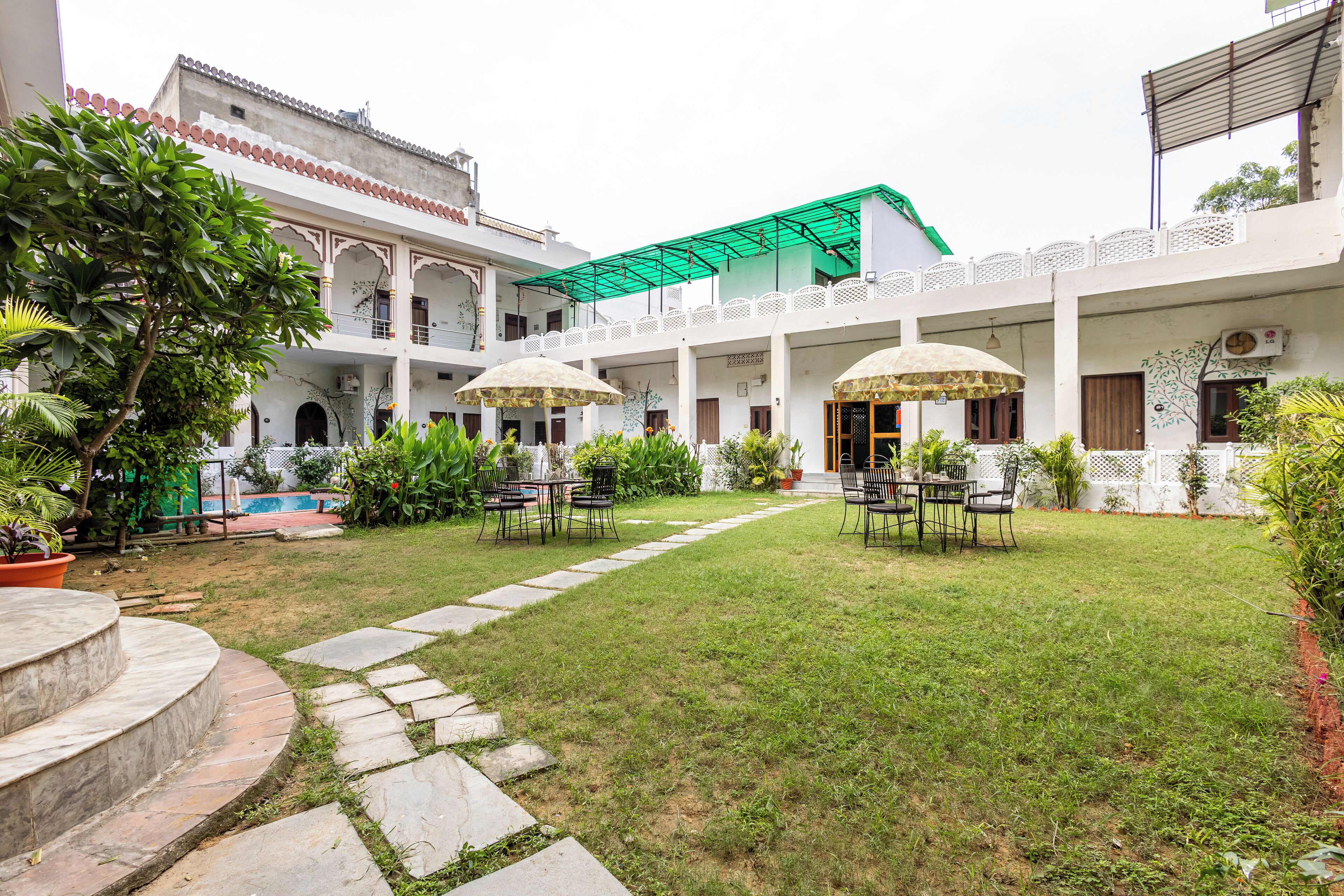 A courtyard with a central lawn, surrounded by white buildings with arches and a green roof. There are outdoor tables and chairs under umbrellas.