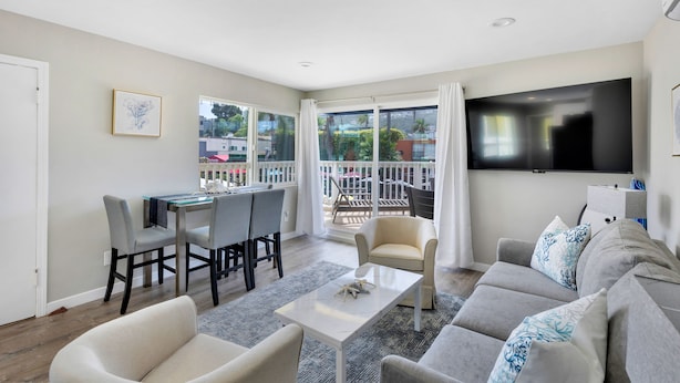 A modern living room with a dining area, a sofa set, a flat-screen TV, and a balcony with a view of the city.