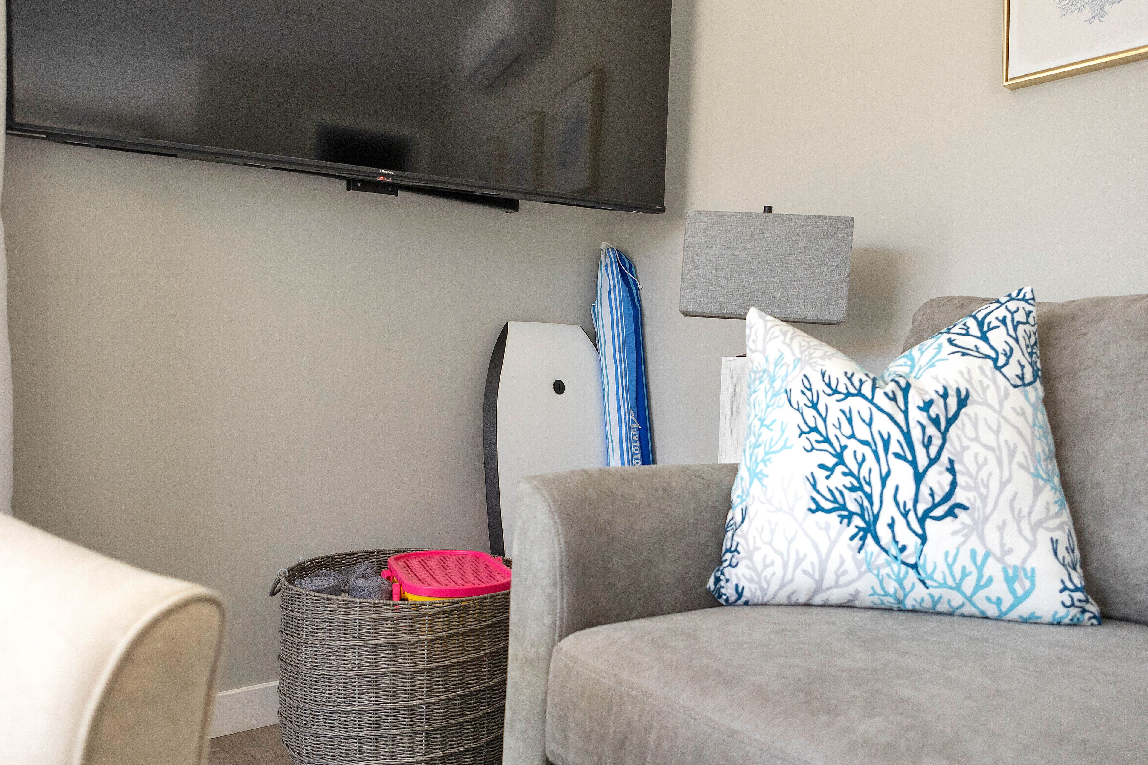 Un salon avec un canapé gris, un téléviseur à écran plat fixé au mur, un panier en osier avec un couvercle rouge et un tableau encadré accroché au mur.