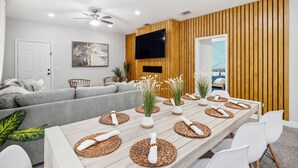 Una sala de estar moderna con una larga mesa de comedor de madera dispuesta con individuales y jarrones, un sofá gris y un televisor de pantalla plana montado en una pared de madera.