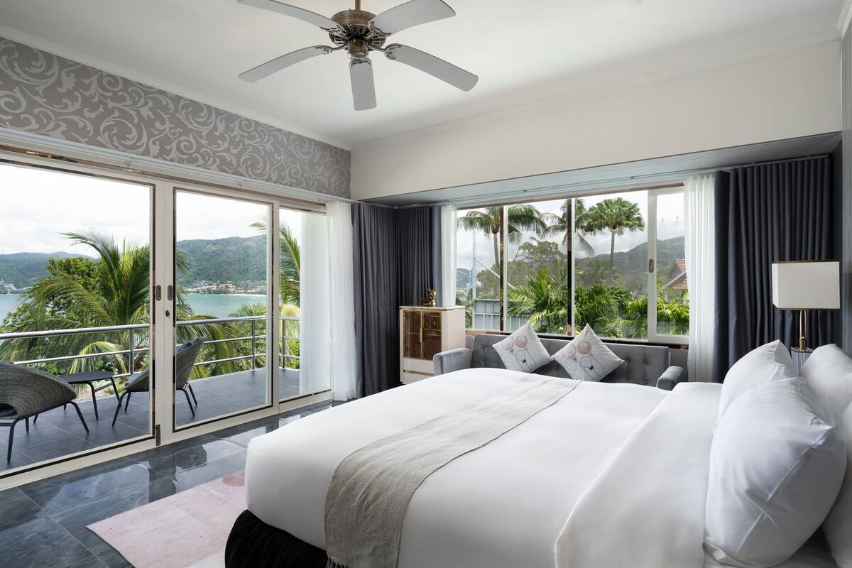 A spacious bedroom with a large bed, a ceiling fan, and a view of the ocean through floor-to-ceiling windows.