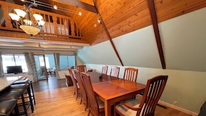 Un comedor con una gran mesa de madera y sillas, una cocina al fondo, y un altillo con vigas de madera y una araña.