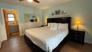 Un dormitorio con una cama grande, lámparas de noche, ventilador de techo, baño con lavamanos y espejo, y un televisor montado en la pared.