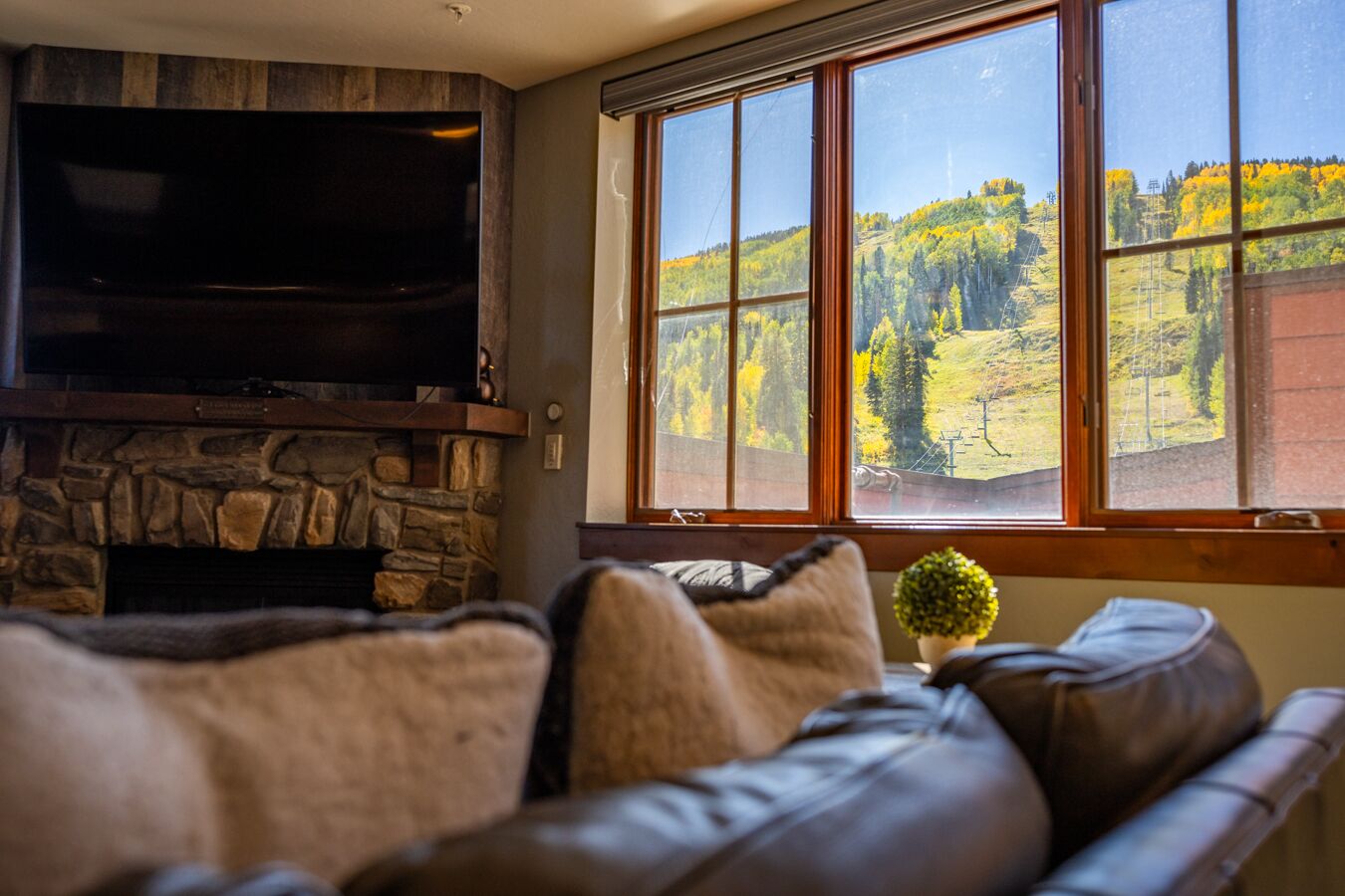 A living room with a stone fireplace, a flat-screen TV mounted above it, and a large window offering a view of a forested hillside.